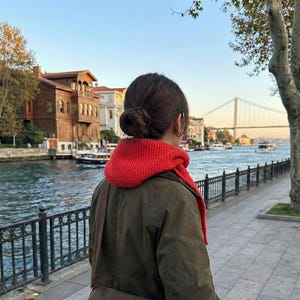 Puede incluir: Una persona con una bufanda roja de punto y una chaqueta verde oliva, de pie cerca de una zona costera con edificios y un puente al fondo. La persona tiene el pelo recogido en un mo&ntilde;o.