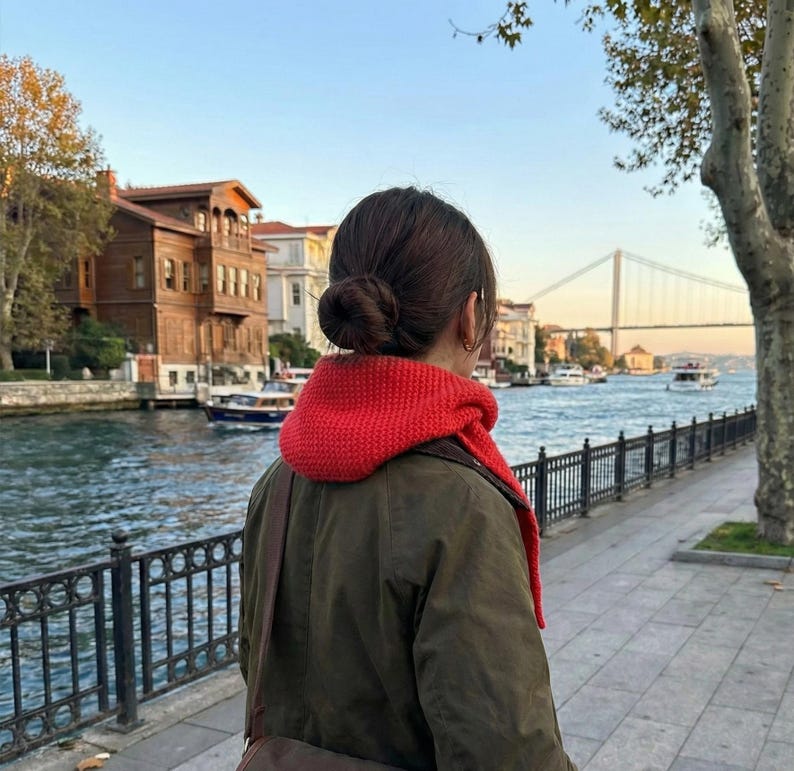 Puede incluir: Una persona con una bufanda roja de punto y una chaqueta verde oliva, de pie cerca de una zona costera con edificios y un puente al fondo. La persona tiene el pelo recogido en un mo&ntilde;o.