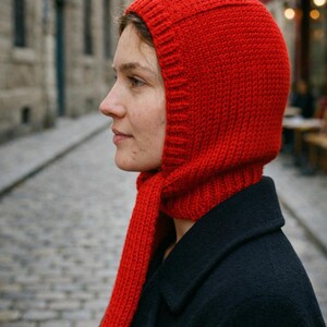 Side profile of a person wearing a bright red knit balabonnet on a cobblestone street with blurred stone buildings and café seating in the background.