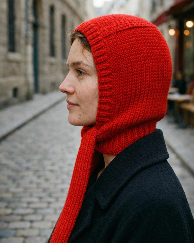 Side profile of a person wearing a bright red knit balabonnet on a cobblestone street with blurred stone buildings and café seating in the background.