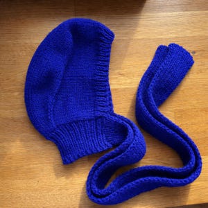 Close-up of a royal blue knit hood laid flat on a wooden floor, highlighting the ribbed neckline and textured stitches; a long knitted tie strap lies beside it with the end folded over.