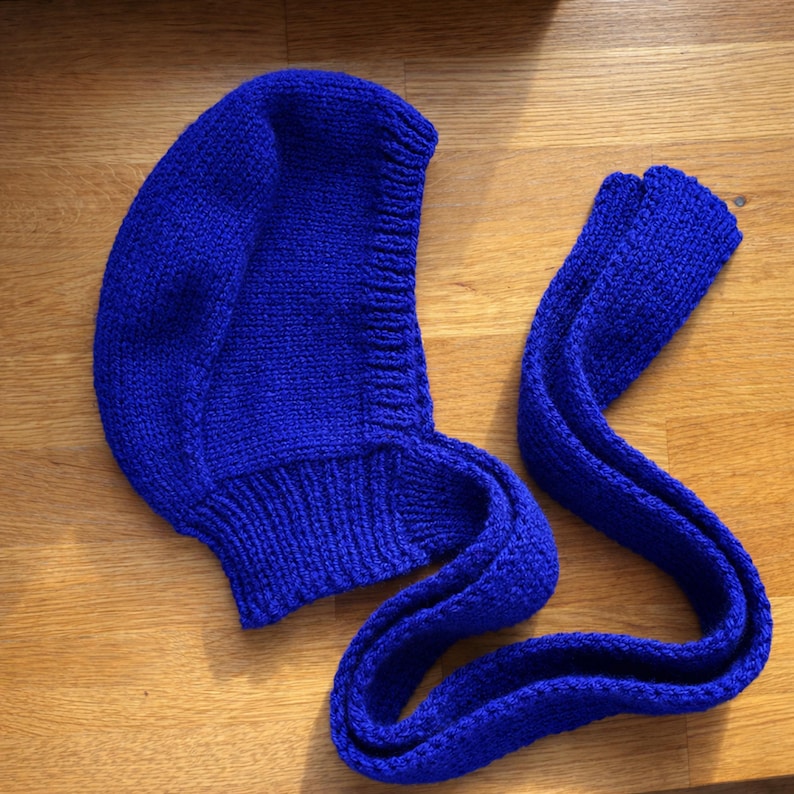 Close-up of a royal blue knit hood laid flat on a wooden floor, highlighting the ribbed neckline and textured stitches; a long knitted tie strap lies beside it with the end folded over.