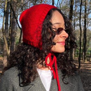 May include: A bright red knit bonnet featuring a textured stitch pattern and long matching red yarn ties that hang down from the chin area. The headwear is constructed from a soft yarn material and is designed to cover the top and sides of the head.