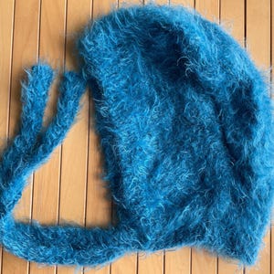 A soft, turquoise fuzzy knitted bonnet lying on a light-colored wooden table. The bonnet features long matching ties and a fluffy, mohair-style texture. Bright, natural sunlight streams across the scene, highlighting the delicate fibers of the knitwear.