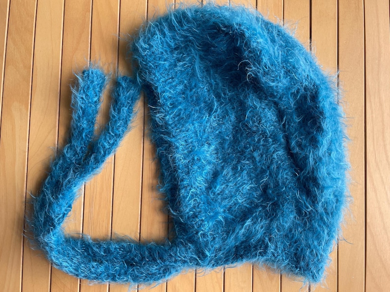 A soft, turquoise fuzzy knitted bonnet lying on a light-colored wooden table. The bonnet features long matching ties and a fluffy, mohair-style texture. Bright, natural sunlight streams across the scene, highlighting the delicate fibers of the knitwear.