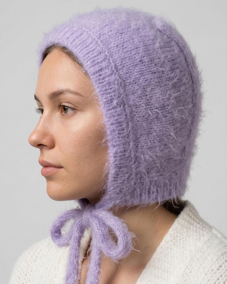 A close-up profile view of a woman wearing a fuzzy, light purple knitted bonnet. The bonnet has a soft, mohair-like texture, a ribbed border around the face, and is tied under the chin with a matching purple knit bow. She is also wearing a chunky white knit sweater against a plain grey background.