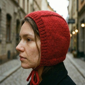 May include: A knitted red bonnet with a rounded crown and a ribbed brim. The bonnet features long ties for fastening under the chin. The hat is made from a thick, textured yarn.