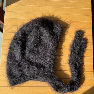 A soft, black fuzzy knitted bonnet lying on a light-colored wooden table. The bonnet features long matching ties and a fluffy, mohair-style texture. Bright, natural sunlight streams across the scene, highlighting the delicate fibers of the knitwear.