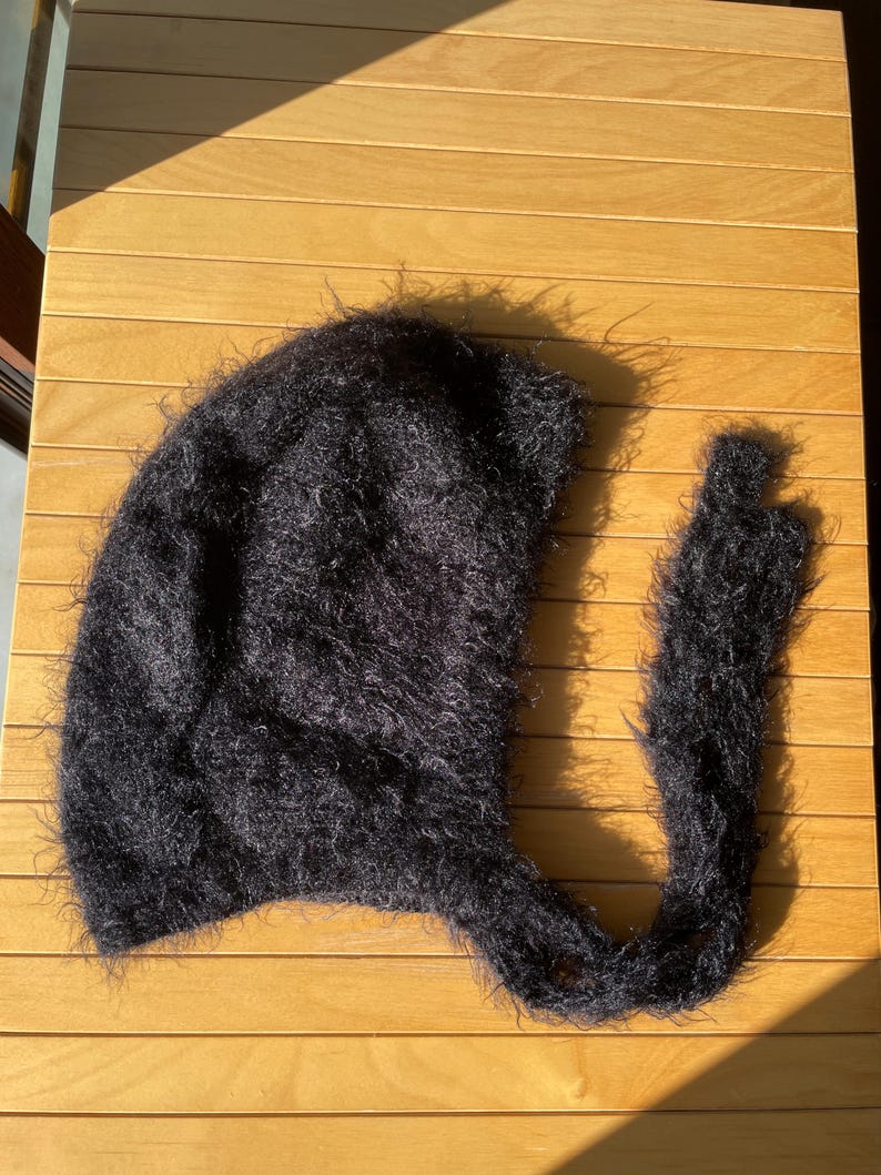 A soft, black fuzzy knitted bonnet lying on a light-colored wooden table. The bonnet features long matching ties and a fluffy, mohair-style texture. Bright, natural sunlight streams across the scene, highlighting the delicate fibers of the knitwear.