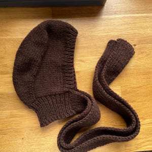 A dark brown knitted balabonnet. The hood is rounded and fits snugly around the face, with a ribbed edge. The scarf is long and narrow, also knitted in a matching dark brown yarn. The set is displayed on a wooden surface.