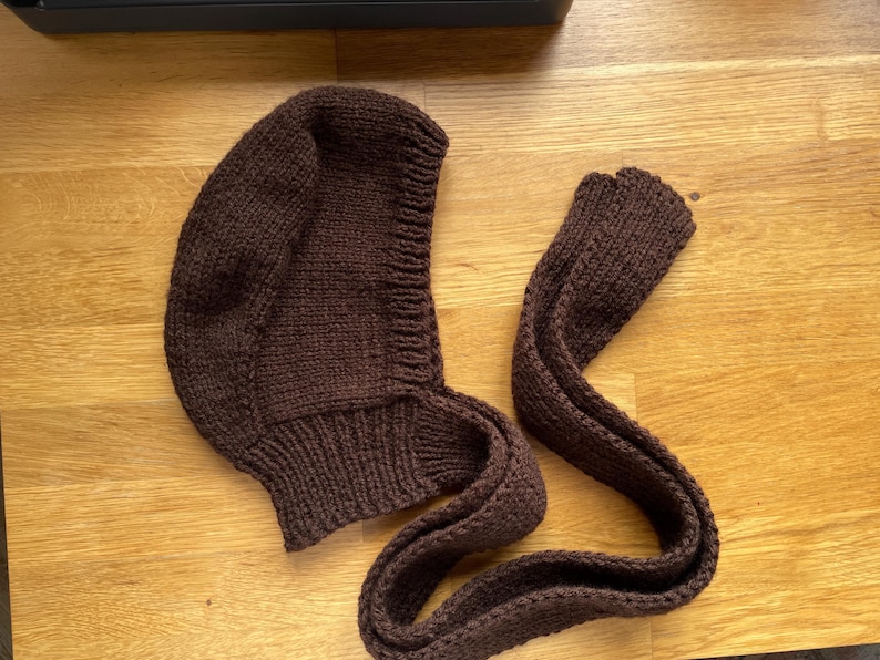 A dark brown knitted balabonnet. The hood is rounded and fits snugly around the face, with a ribbed edge. The scarf is long and narrow, also knitted in a matching dark brown yarn. The set is displayed on a wooden surface.