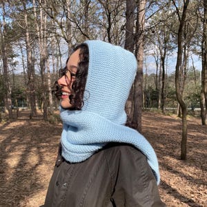 May include: A light blue knitted hood with an integrated scarf, featuring a textured garter stitch pattern. The hood covers the head and neck, with the scarf portion draping over the shoulders, worn over a dark olive green jacket.