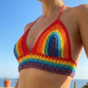 A woman wearing a vibrant, handmade rainbow crochet bikini top against a clear blue sky and ocean backdrop. The halter-style top features concentric triangular patterns in red, orange, yellow, green, blue, and purple. The photo is taken from a side angle, showcasing the textured stitching and the colorful striped band at the bottom.