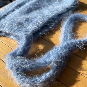 A soft, blue fuzzy knitted bonnet lying on a light-colored wooden table. The bonnet features long matching ties and a fluffy, mohair-style texture. Bright, natural sunlight streams across the scene, highlighting the delicate fibers of the knitwear.
