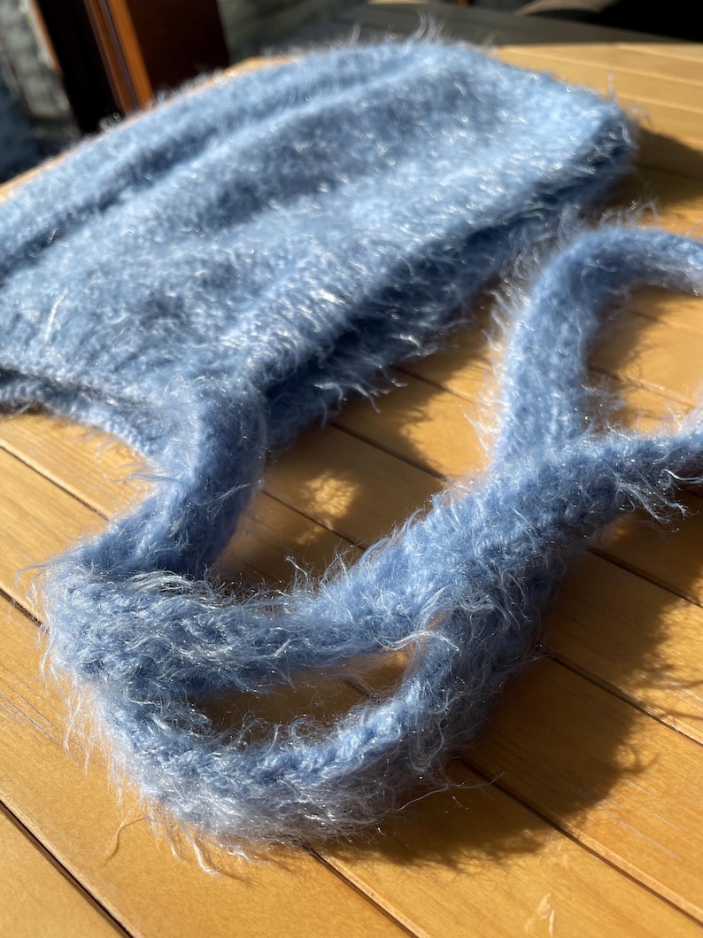 A soft, blue fuzzy knitted bonnet lying on a light-colored wooden table. The bonnet features long matching ties and a fluffy, mohair-style texture. Bright, natural sunlight streams across the scene, highlighting the delicate fibers of the knitwear.