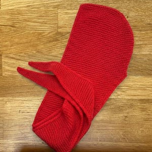 Puede incluir: Una capucha roja brillante tejida con extremos de bufanda, exhibida sobre una superficie de madera. La capucha es de un solo color y parece estar hecha de un hilo grueso, adecuada para el clima fr&iacute;o.