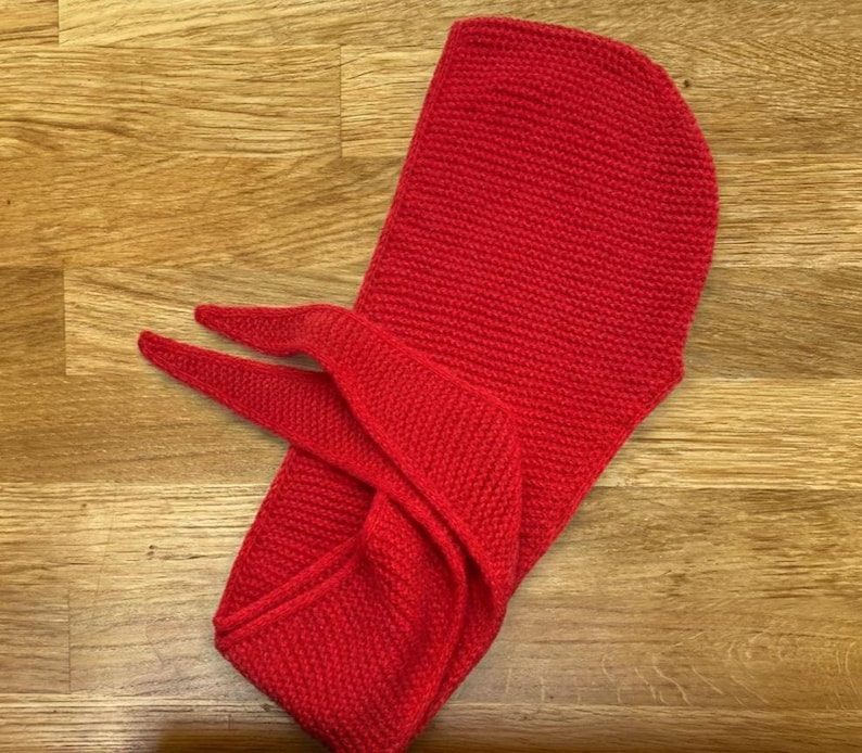 Puede incluir: Una capucha roja brillante tejida con extremos de bufanda, exhibida sobre una superficie de madera. La capucha es de un solo color y parece estar hecha de un hilo grueso, adecuada para el clima fr&iacute;o.