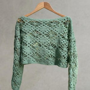 A light green long-sleeved crochet crop top displayed on a wooden hanger against a neutral wall, highlighting the symmetrical floral knit pattern and scale.