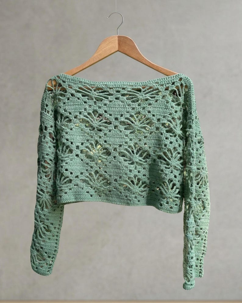 A light green long-sleeved crochet crop top displayed on a wooden hanger against a neutral wall, highlighting the symmetrical floral knit pattern and scale.