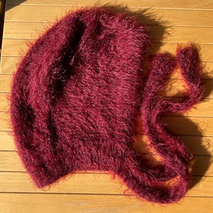 A soft, burgundy fuzzy knitted bonnet lying on a light-colored wooden table. The bonnet features long matching ties and a fluffy, mohair-style texture. Bright, natural sunlight streams across the scene, highlighting the delicate fibers of the knitwear.