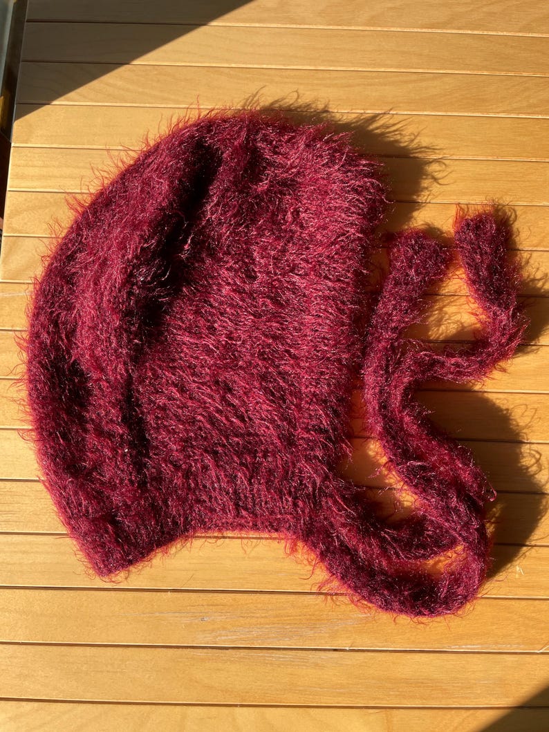 A soft, burgundy fuzzy knitted bonnet lying on a light-colored wooden table. The bonnet features long matching ties and a fluffy, mohair-style texture. Bright, natural sunlight streams across the scene, highlighting the delicate fibers of the knitwear.