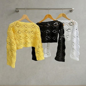 Three long-sleeved crochet crop tops in yellow, black, and white hang on wooden hangers from a metal rail. Each top features an intricate, open-knit floral star pattern with a scalloped texture. The background is a neutral grey wall.