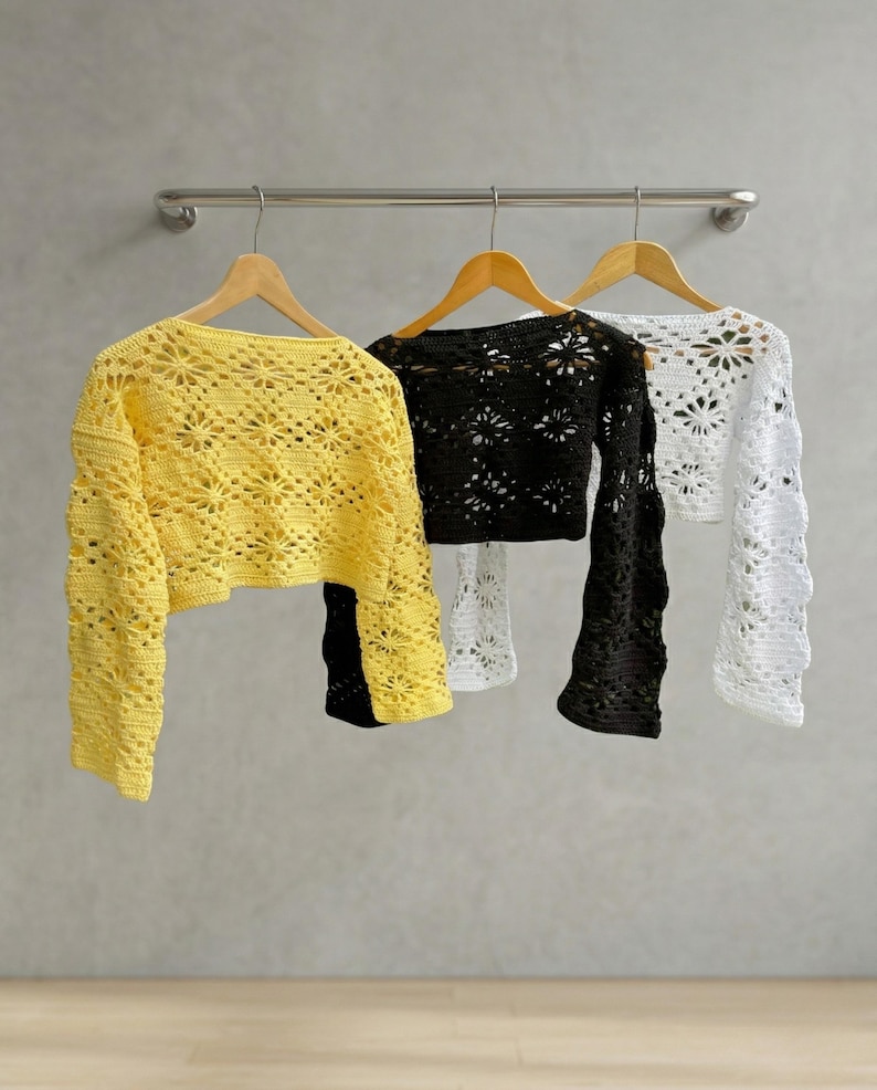 Three long-sleeved crochet crop tops in yellow, black, and white hang on wooden hangers from a metal rail. Each top features an intricate, open-knit floral star pattern with a scalloped texture. The background is a neutral grey wall.