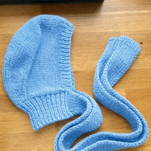 Light blue knit hood laid flat on a wooden floor, showing the curved head shape and ribbed neckline; a long knitted tie strap is extended beside it with the end folded over.