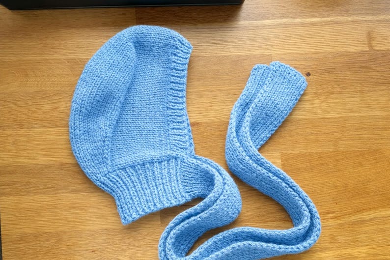 Light blue knit hood laid flat on a wooden floor, showing the curved head shape and ribbed neckline; a long knitted tie strap is extended beside it with the end folded over.