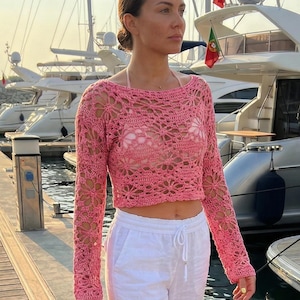 A model standing on a pier in front of yachts wearing a vibrant pink floral crochet crop top with long sleeves, showing the delicate open-knit texture.