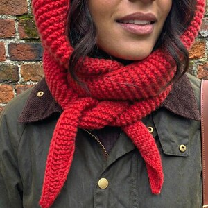 Puede incluir: Conjunto de capucha y bufanda de punto rojo vibrante. La capucha enmarca la cara, mientras que la bufanda est&aacute; anudada al cuello. El conjunto se lleva sobre una chaqueta verde oscuro con cuello de pana marr&oacute;n y botones dorados. Se ve una correa de cuero marr&oacute;n.