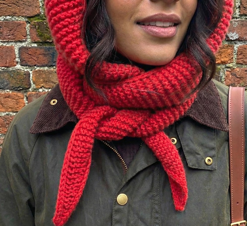 Puede incluir: Conjunto de capucha y bufanda de punto rojo vibrante. La capucha enmarca la cara, mientras que la bufanda est&aacute; anudada al cuello. El conjunto se lleva sobre una chaqueta verde oscuro con cuello de pana marr&oacute;n y botones dorados. Se ve una correa de cuero marr&oacute;n.