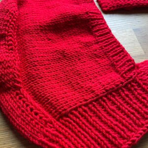 May include: A bright red hand-knitted bonnet featuring a textured stitch pattern and a ribbed border along the edge. The garment is constructed with a dense knit structure and includes a separate matching red knitted piece positioned nearby.