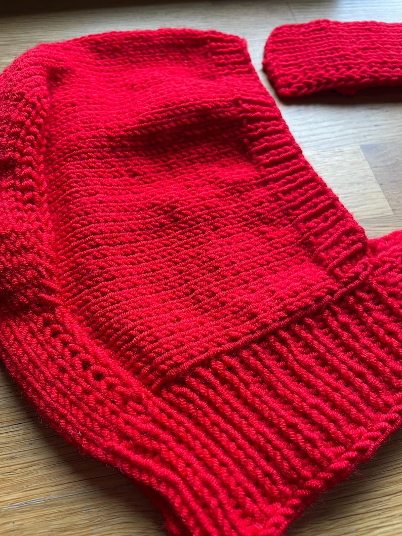 May include: A bright red hand-knitted bonnet featuring a textured stitch pattern and a ribbed border along the edge. The garment is constructed with a dense knit structure and includes a separate matching red knitted piece positioned nearby.