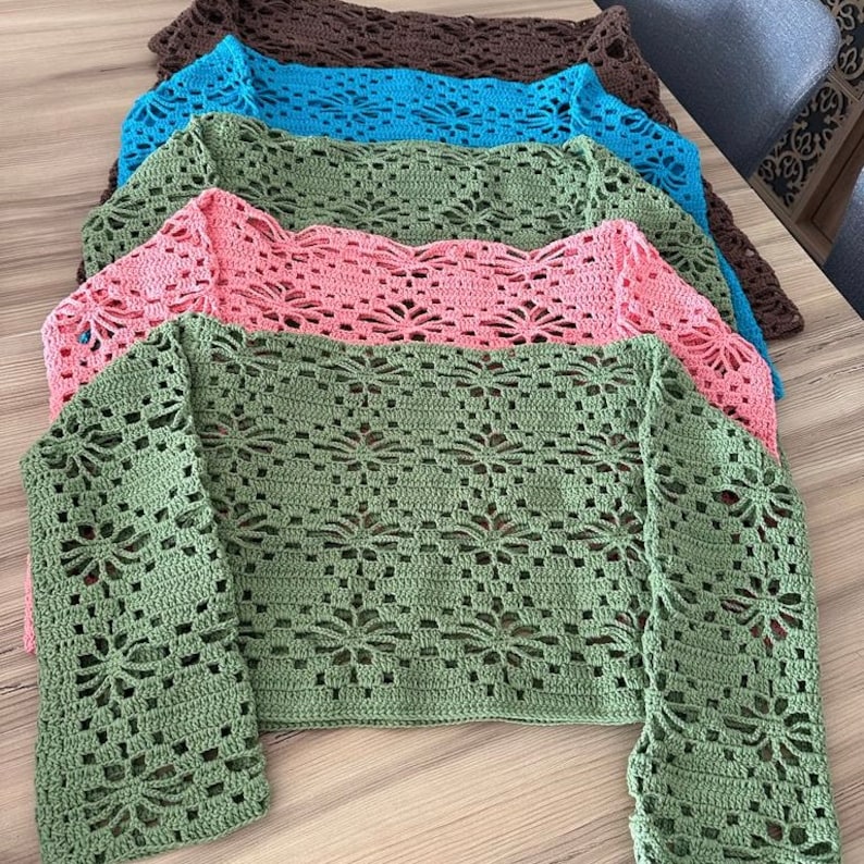 A stack of four crocheted tops in different colors, including green, pink, blue, and brown. The tops have a delicate, lacy pattern and are perfect for layering or wearing on their own.