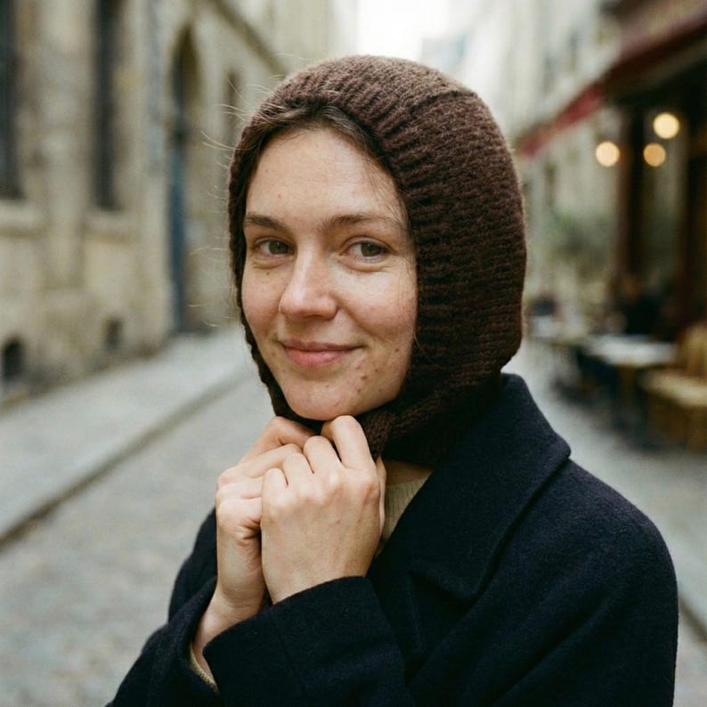 Close-up of a person wearing a dark brown ribbed knit balabonnet, holding the ties under the chin, with a black coat visible and a blurred cobblestone street and café lights in the background.