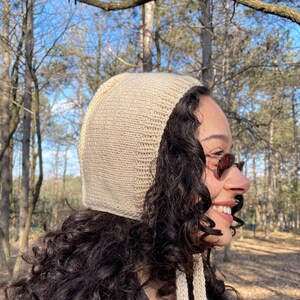 May include: A beige knitted hood with long ties. The hood is made of a soft, textured yarn and is worn over dark curly hair. The background features a forest with trees and sunlight.