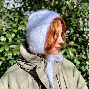 May include: A white fuzzy knit bonnet with a soft, textured surface and long matching ties that fasten under the chin. The headwear features a close-fitting design crafted from a lightweight, airy yarn that provides a gentle, halo-like appearance.