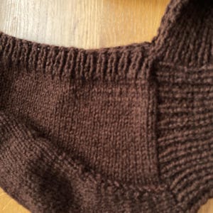 A close-up of a brown knitted balabonnet. The garment has a ribbed neckline and a textured, woven appearance. The color is a deep, warm brown.