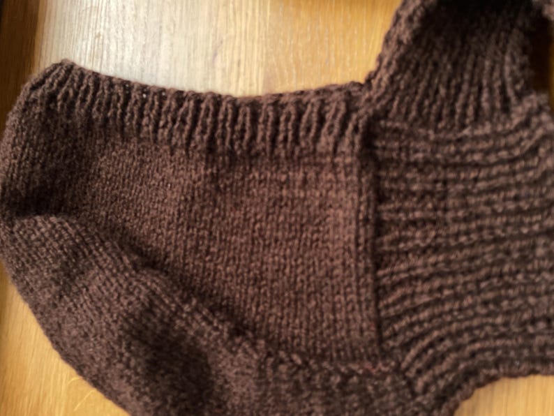 A close-up of a brown knitted balabonnet. The garment has a ribbed neckline and a textured, woven appearance. The color is a deep, warm brown.