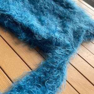 A soft, turquoise fuzzy knitted bonnet lying on a light-colored wooden table. The bonnet features long matching ties and a fluffy, mohair-style texture. Bright, natural sunlight streams across the scene, highlighting the delicate fibers of the knitwear.