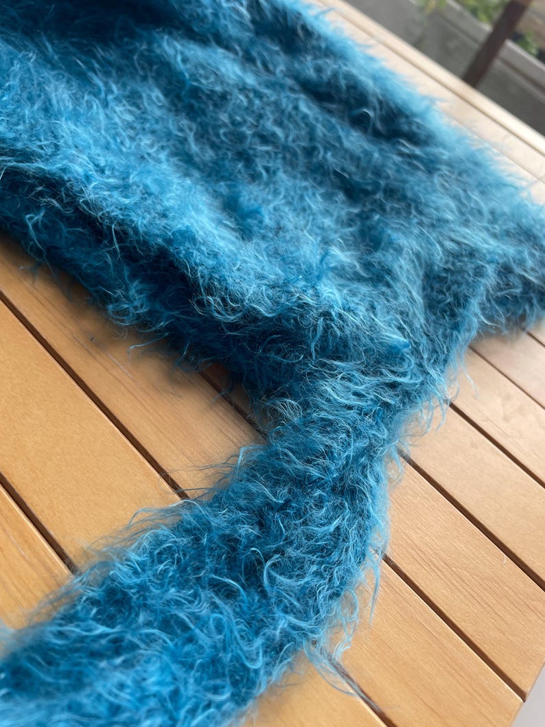 A soft, turquoise fuzzy knitted bonnet lying on a light-colored wooden table. The bonnet features long matching ties and a fluffy, mohair-style texture. Bright, natural sunlight streams across the scene, highlighting the delicate fibers of the knitwear.