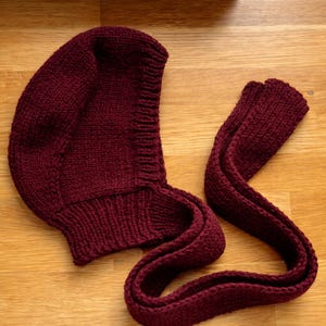Burgundy knit hood laid flat on a wooden floor, showing the curved hood shape and ribbed neckline; a long knitted tie strap is extended beside it with the end folded over.