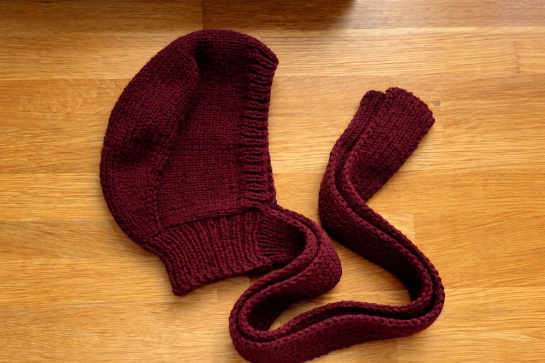 Burgundy knit hood laid flat on a wooden floor, showing the curved hood shape and ribbed neckline; a long knitted tie strap is extended beside it with the end folded over.