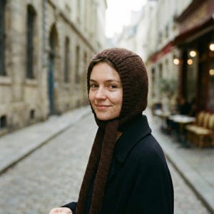 Three-quarter view of a person on a cobblestone street wearing a dark brown ribbed knit balabonnet with long ties draped down the front, black coat visible; blurred stone buildings and café seating behind.