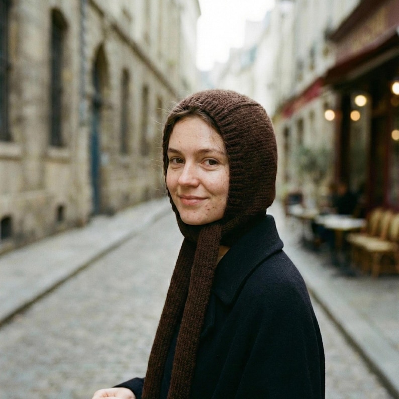 Three-quarter view of a person on a cobblestone street wearing a dark brown ribbed knit balabonnet with long ties draped down the front, black coat visible; blurred stone buildings and café seating behind.
