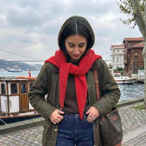 Puede incluir: Una persona lleva una bufanda roja de punto, una chaqueta verde oscuro y vaqueros azules. Un bolso de cuero marr&oacute;n cuelga de su hombro. El fondo incluye agua, barcos y edificios bajo un cielo nublado.