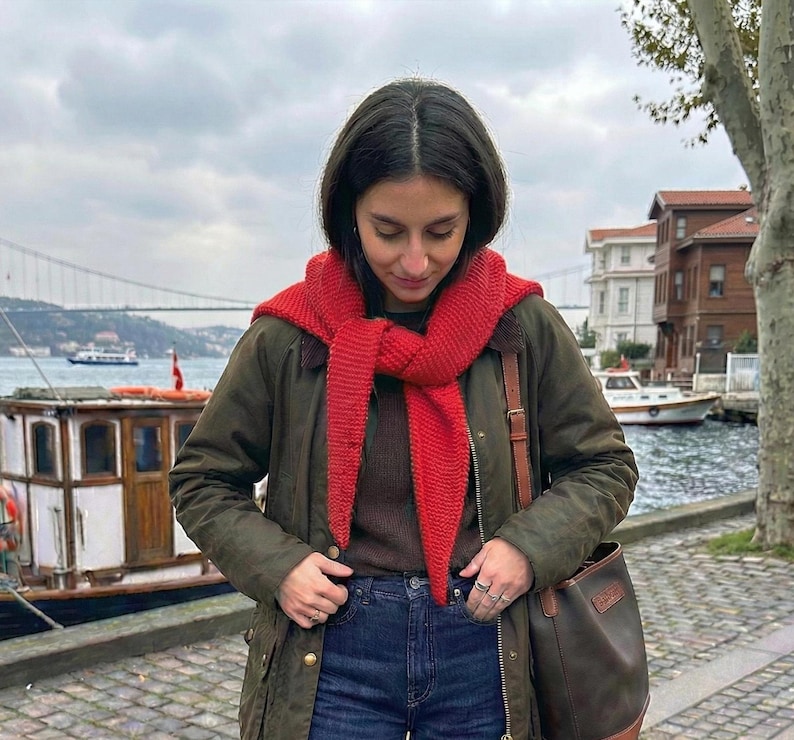 Puede incluir: Una persona lleva una bufanda roja de punto, una chaqueta verde oscuro y vaqueros azules. Un bolso de cuero marr&oacute;n cuelga de su hombro. El fondo incluye agua, barcos y edificios bajo un cielo nublado.