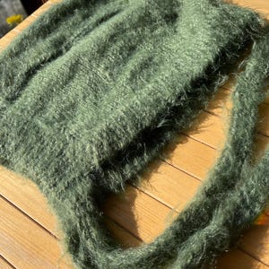 A soft, green fuzzy knitted bonnet lying on a light-colored wooden table. The bonnet features long matching ties and a fluffy, mohair-style texture. Bright, natural sunlight streams across the scene, highlighting the delicate fibers of the knitwear.