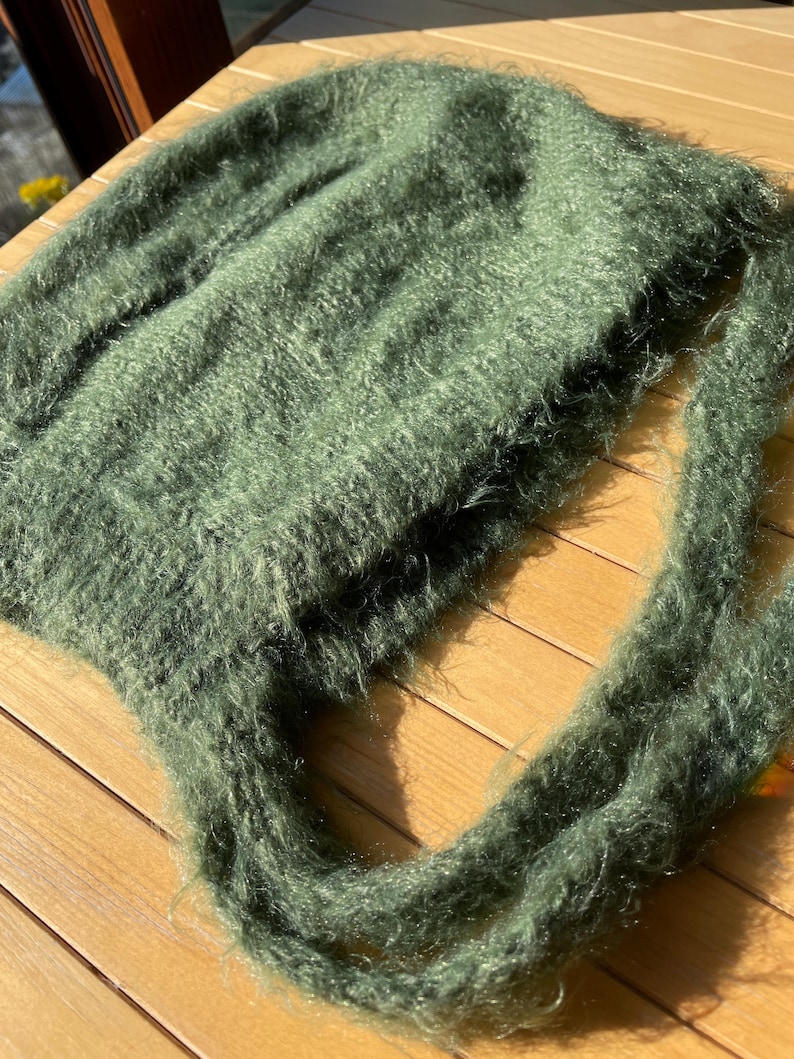 A soft, green fuzzy knitted bonnet lying on a light-colored wooden table. The bonnet features long matching ties and a fluffy, mohair-style texture. Bright, natural sunlight streams across the scene, highlighting the delicate fibers of the knitwear.